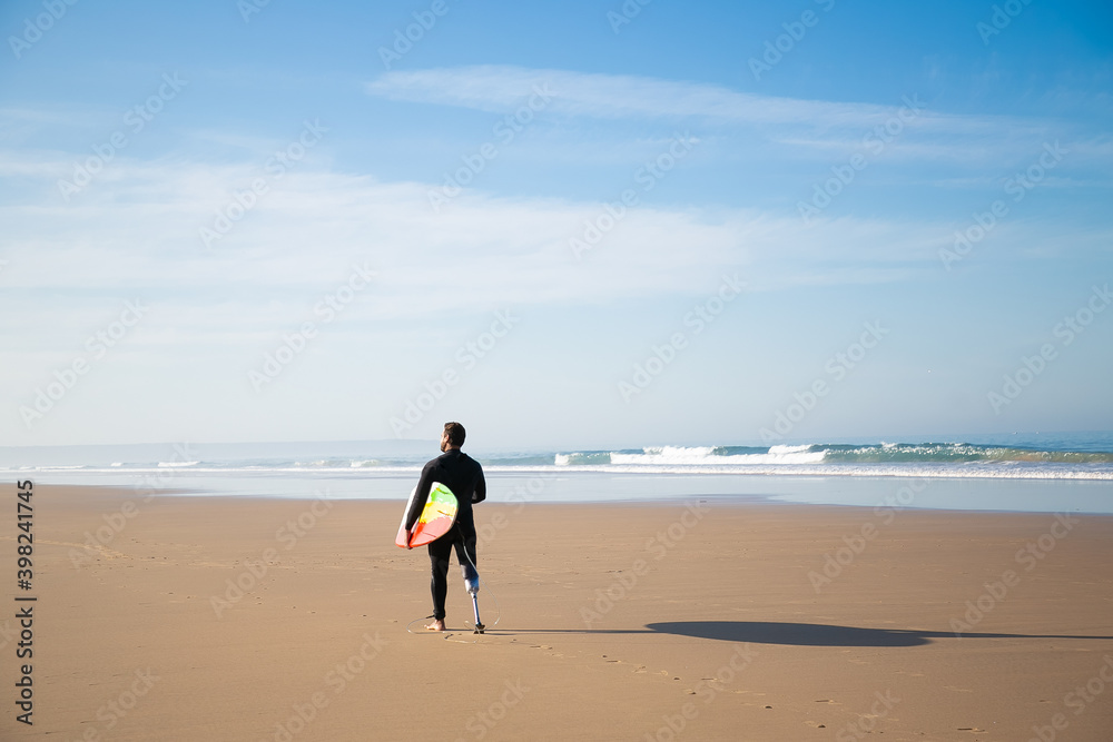 Back view of surfer standing on sand beach with board. Disabled active amputee in black wetsuit walking along sea cast during sunny day. Physical disability, surfing and extreme sport concept
