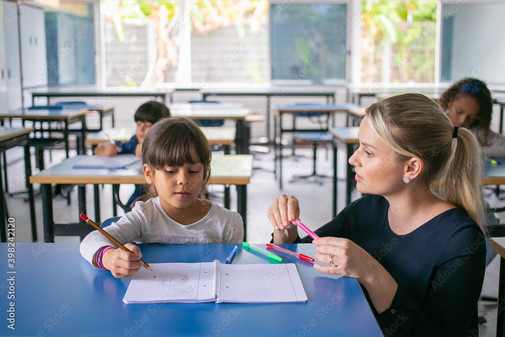 Fototapeta premium Serious primary school teacher helping girl to cope with her task. Schoolgirl drawing in her copybook in class. Education or back to school concept