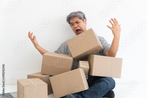 Asian Shopaholic man Shocked and sitting on the floor in the living room and Cardboard Box on top of him after The courier delivered to the home. Concept of shopping online and Shopaholic.