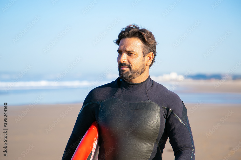 Portrait of handsome surfer standing on beach with surfboard and looking away. Pensive Caucasian middle-aged man holding surfboard and thinking about something. Lifestyle and summer activity concept Portrait of handsome surfer standing on beach with surfboard and looking away. Pensive Caucasian middle-aged man holding surfboard and thinking about something. Lifestyle and summer activity concept