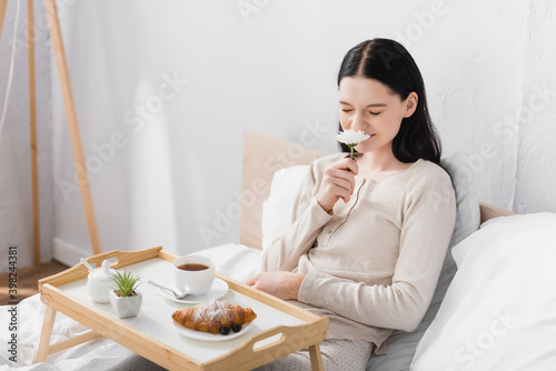 young brunette woman with vitiligo smelling flower near breakfast on tray