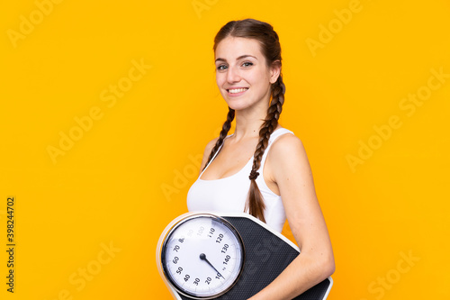 Young woman over isolated yellow background with weighing machine