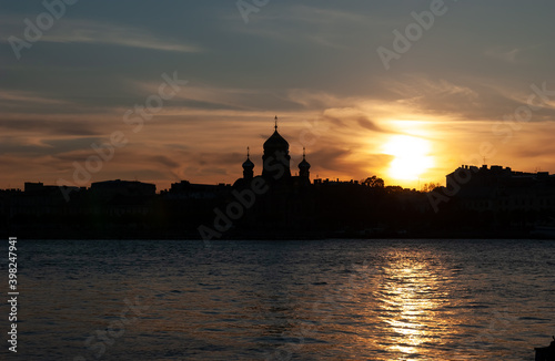 Against the background of a yellow sunset, the contours of the temple on the opposite bank of the Neva River. The sky is painted with sun strokes. Sunny path on dark water. St Petersburg, Russia.