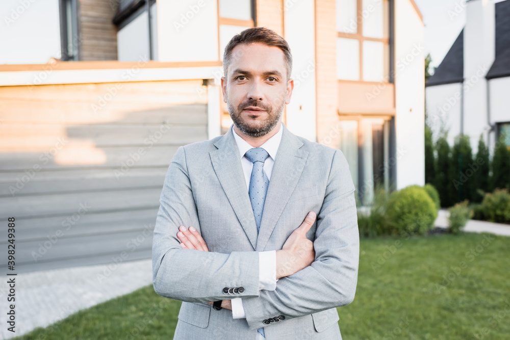 Confident broker with crossed arms looking at camera with blurred houses on background Confident broker with crossed arms looking at camera with blurred houses on background