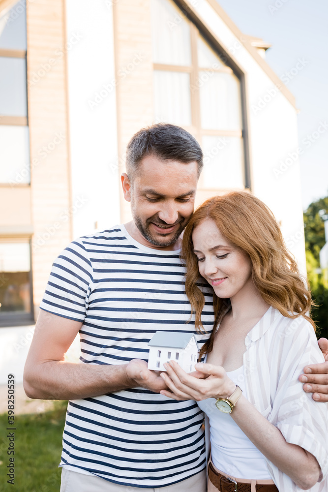 Smiling couple hugging and looking at statuette of house on blurred background Smiling couple hugging and looking at statuette of house on blurred background