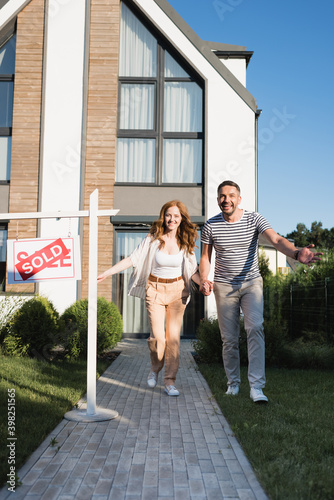 Cheerful couple holding hands while running forward near sign with sold lettering and modern house © LIGHTFIELD STUDIOS