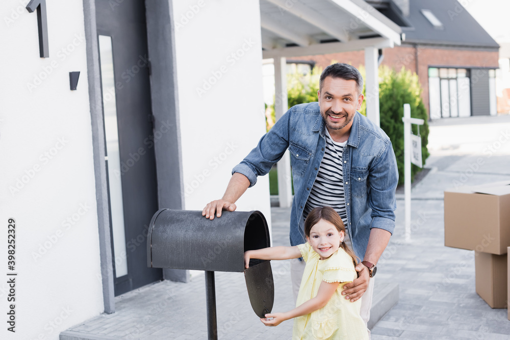 Obraz premium Cheerful father hugging daughter near empty mailbox while looking at camera near house on blurred background