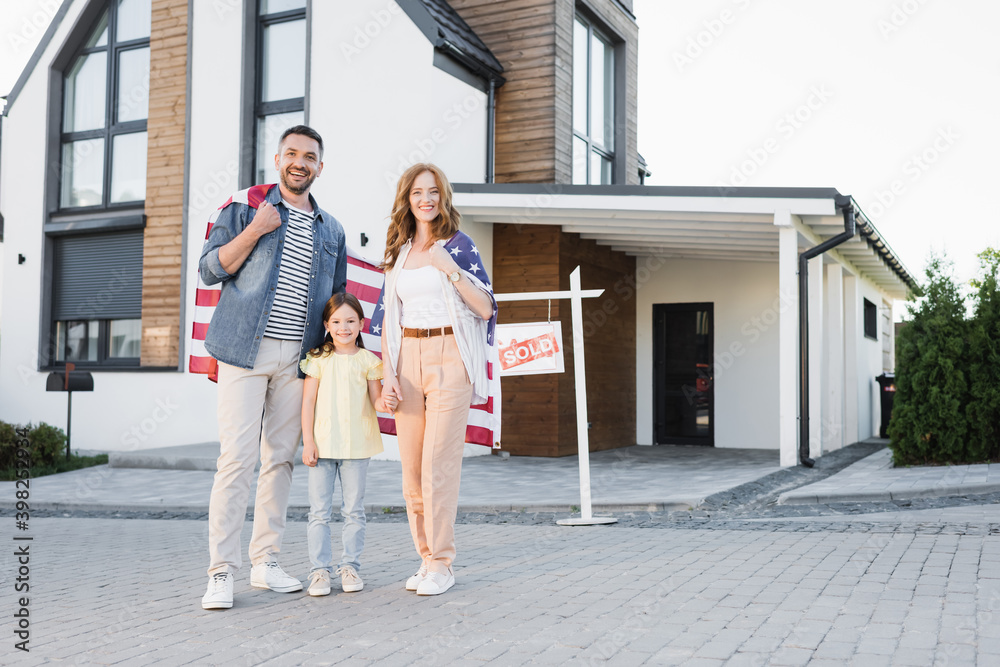 Full length of happy daughter with mom and dad holding american flag while standing together and looking at camera near house