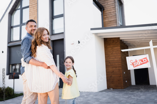 Happy family with daughter hugging and looking at camera near sign with sold lettering and house photo