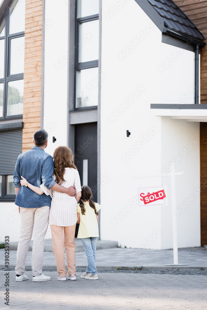 Full length of daughter standing near father and mother hugging near house and sign with sold lettering