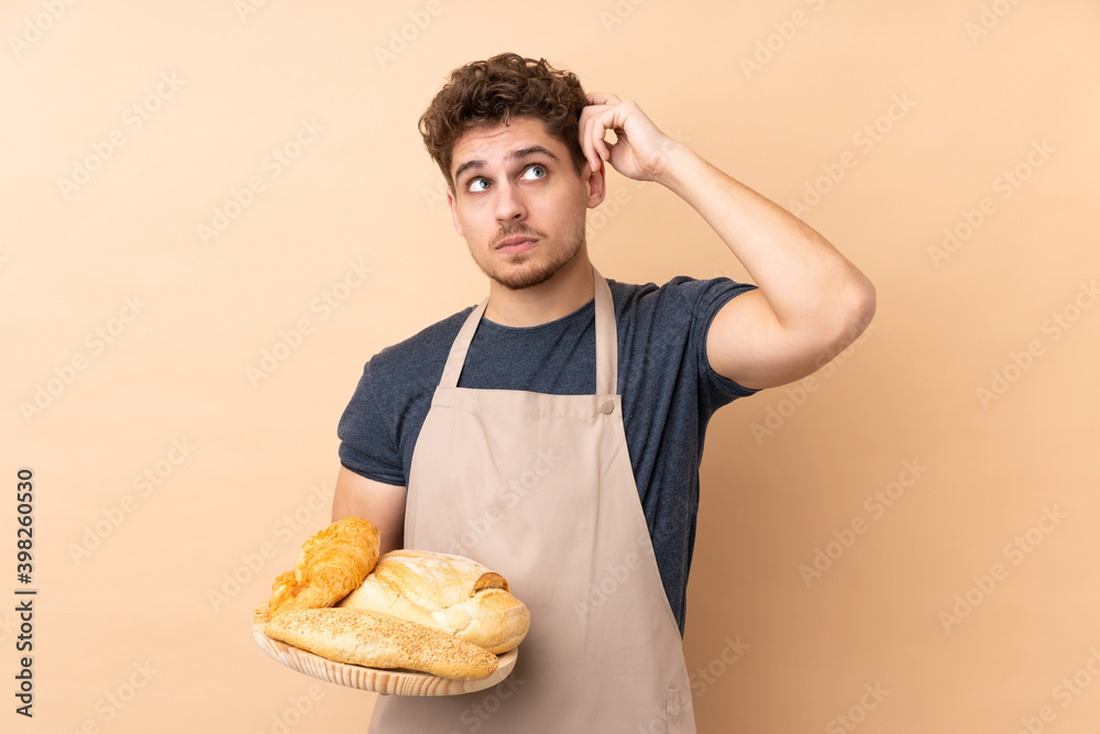 Male baker holding a table with several breads isolated on beige background having doubts and with confuse face expression Male baker holding a table with several breads isolated on beige background having doubts and with confuse face expression