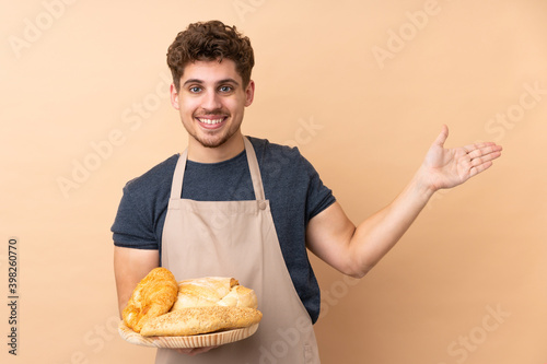 Male baker holding a table with several breads isolated on beige background extending hands to the side for inviting to come