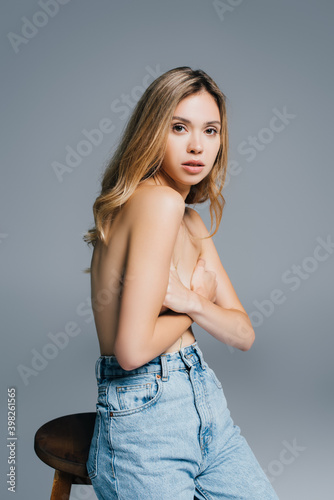 sexy topless woman in jeans covering breasts with hands while looking at camera isolated on grey