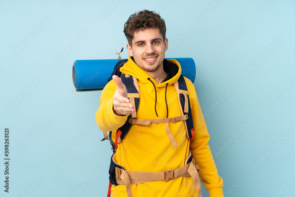 Young mountaineer man with a big backpack isolated on blue  background handshaking after good deal