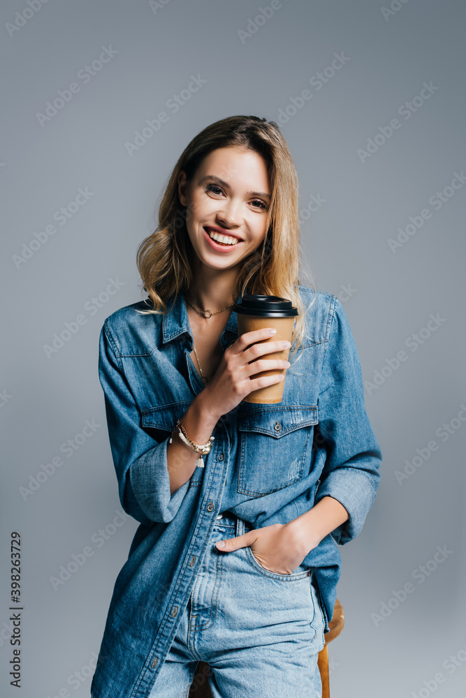 smiling young woman in denim clothes holding hand in pocket and coffee to go isolated on grey smiling young woman in denim clothes holding hand in pocket and coffee to go isolated on grey