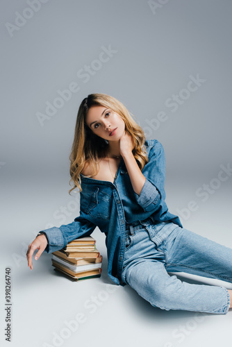 seductive woman in denim shirt and jeans, with naked shoulder, leaning on books while sitting on grey