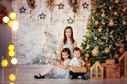 A boy and two girls sit on the floor with gifts against a background of garlands of lights. Three children with gifts at the Christmas tree © Elena 