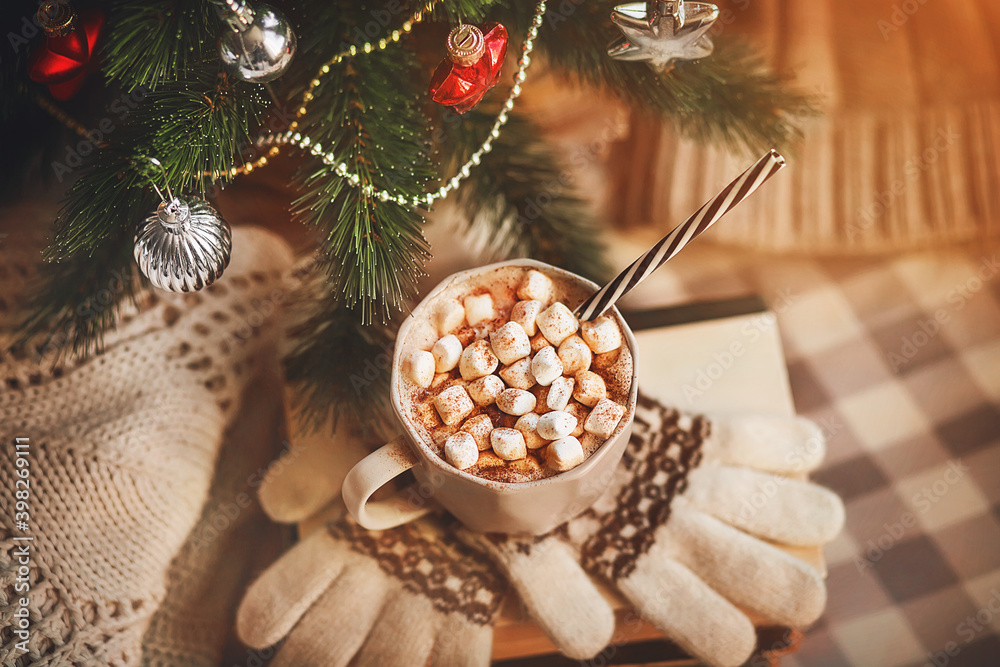 A cup of hot cocoa with marshmallows and cinnamon sits on a stack of books and gloves near a Christmas tree that is decked out with toys, and winter clothes are next to it. New Year. Festive mood. A cup of hot cocoa with marshmallows and cinnamon sits on a stack of books and gloves near a Christmas tree that is decked out with toys, and winter clothes are next to it. New Year. Festive mood.