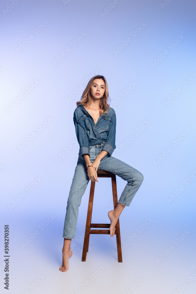 young barefoot woman in denim clothes sitting on high stool on blue young barefoot woman in denim clothes sitting on high stool on blue
