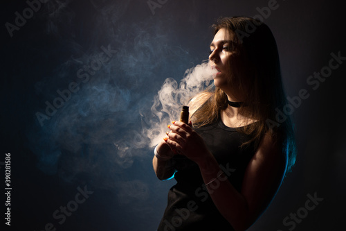 Girl smokes VAPE. Vaping girl. Vaper woman in a cloud of smoke. The girl is holding an electronic cigarette on a dark background. VAPE shop. Vaping concept with space for text.