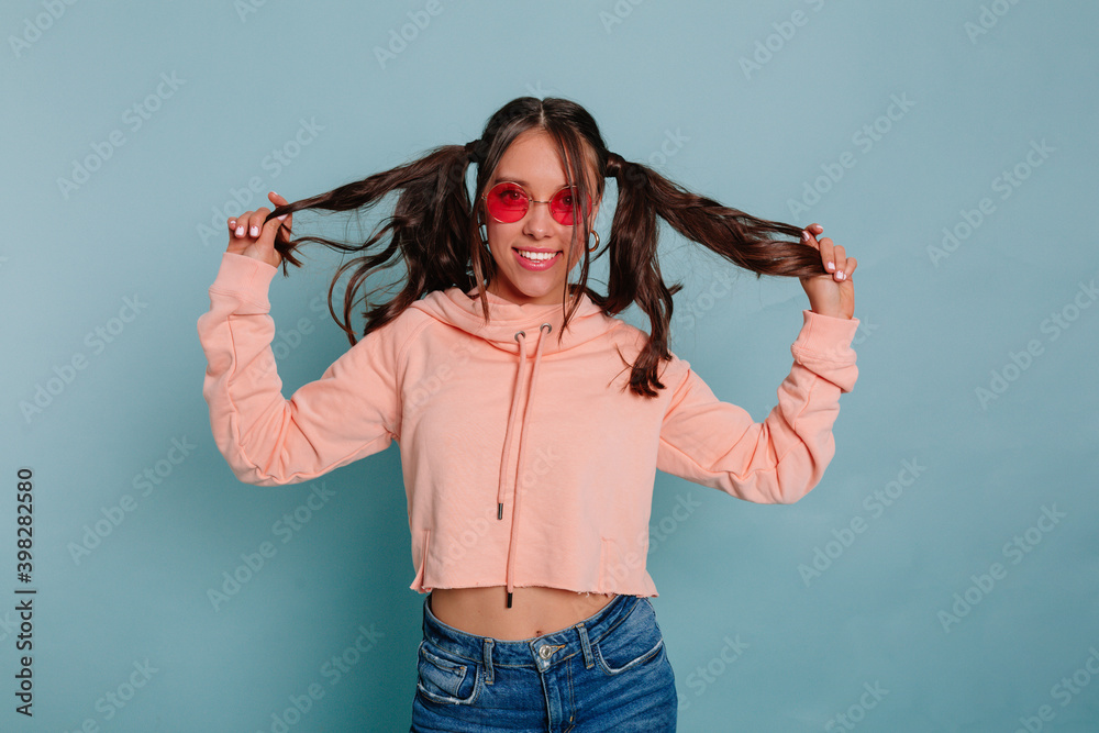 Funny attractive woman with collected hair wearing pink pullover playing with her hair wearing casual clothes posing over blue background