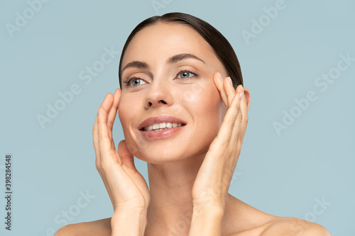 Studio portrait of young woman with natural makeup, combed hair, touching well-groomed pure skin on face, isolated on blue background. Beauty, facelift, anti-aging cosmetic procedure. Banner.  photo