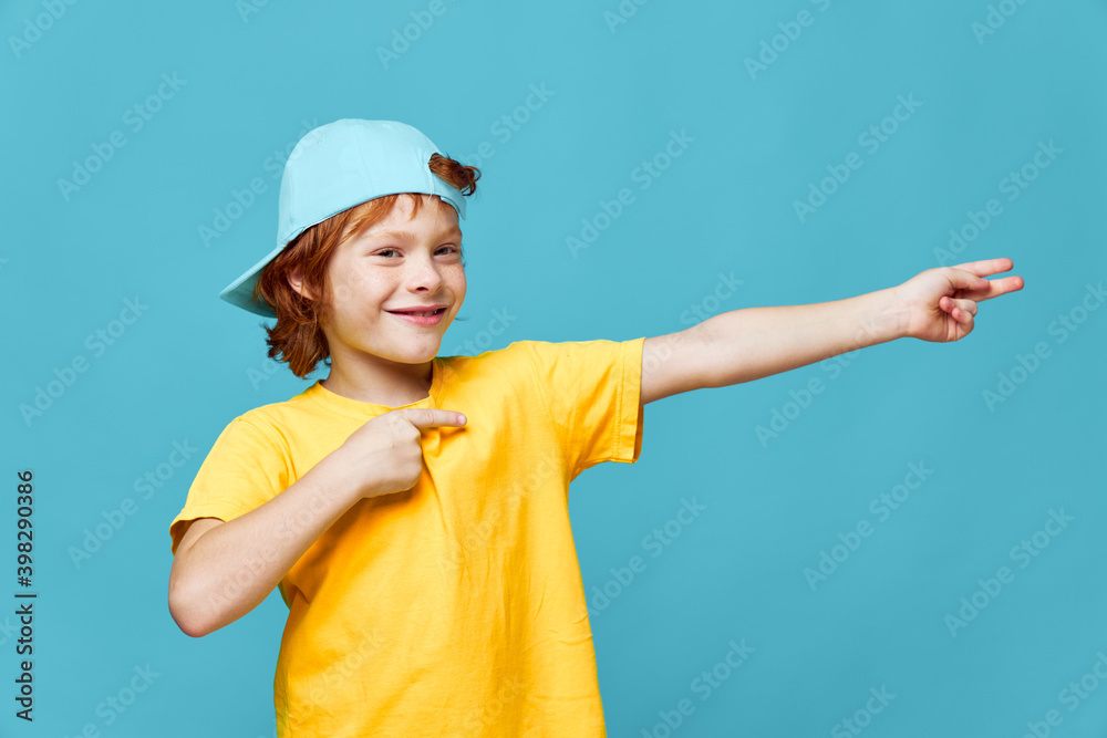 Cheerful redhead child shows fingers to the side smile blue cap yellow t-shirt 