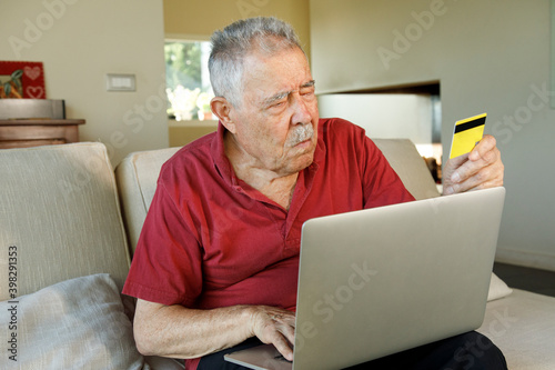 signore anziano con una polo sgargiante è seduto nel divano con un portatile e una carta di credito è intento a fare un ordinazione su internet