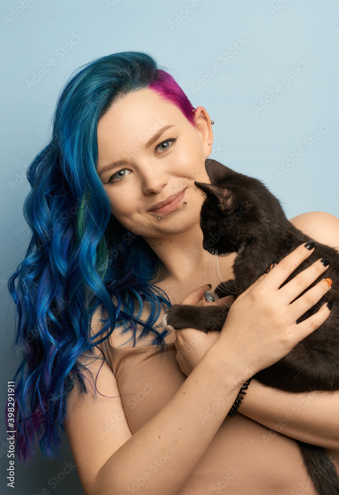 Obraz premium Girl with creative blue coloring and a rainbow in her hair on a blue background holds black cat. Modern minimalistic bright photography for advertising and social networks