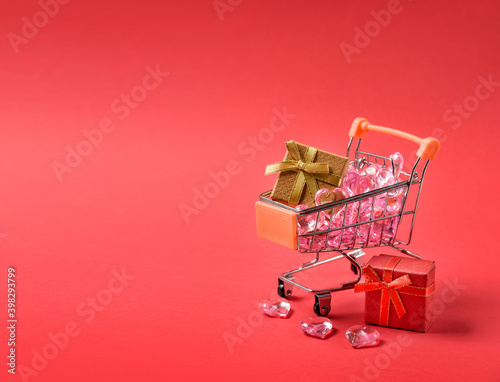 Shopping cart with gifts and glass hearts on a red background with copy space.