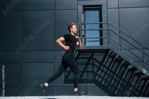 Running near black building. Sportive young guy in black shirt and pants outdoors at daytime