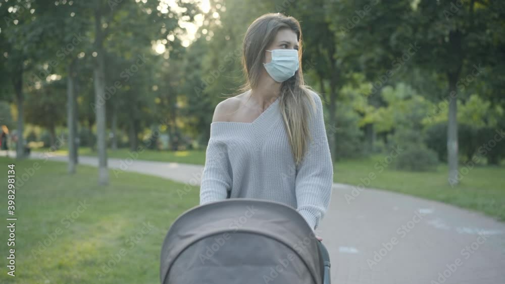 Confident young mother in Covid face mask walking in sunlight with baby