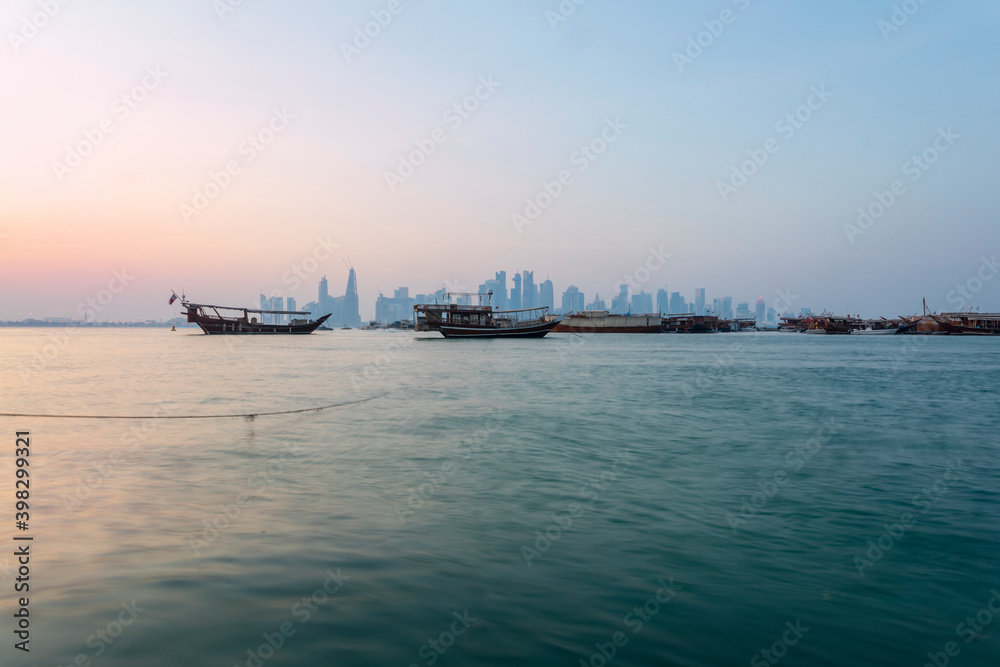 Doha skyline behind some boats on the sea during a beautiful sunset near the port. You can see some boats, buildings, a rope, the sky and the water.