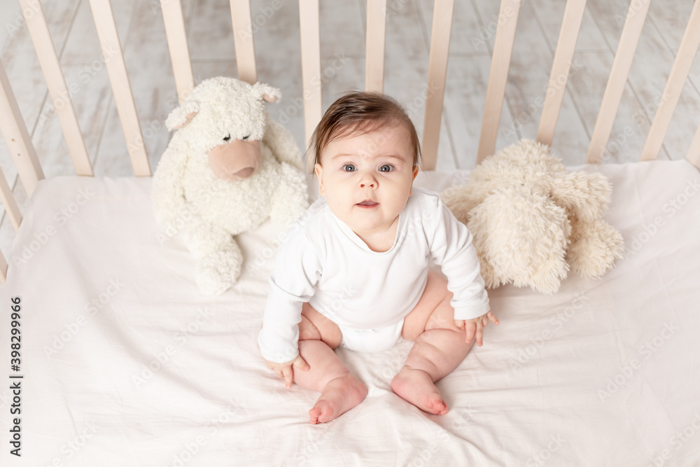 baby six months sitting in a crib in a white bodysuit with toys Teddy bears