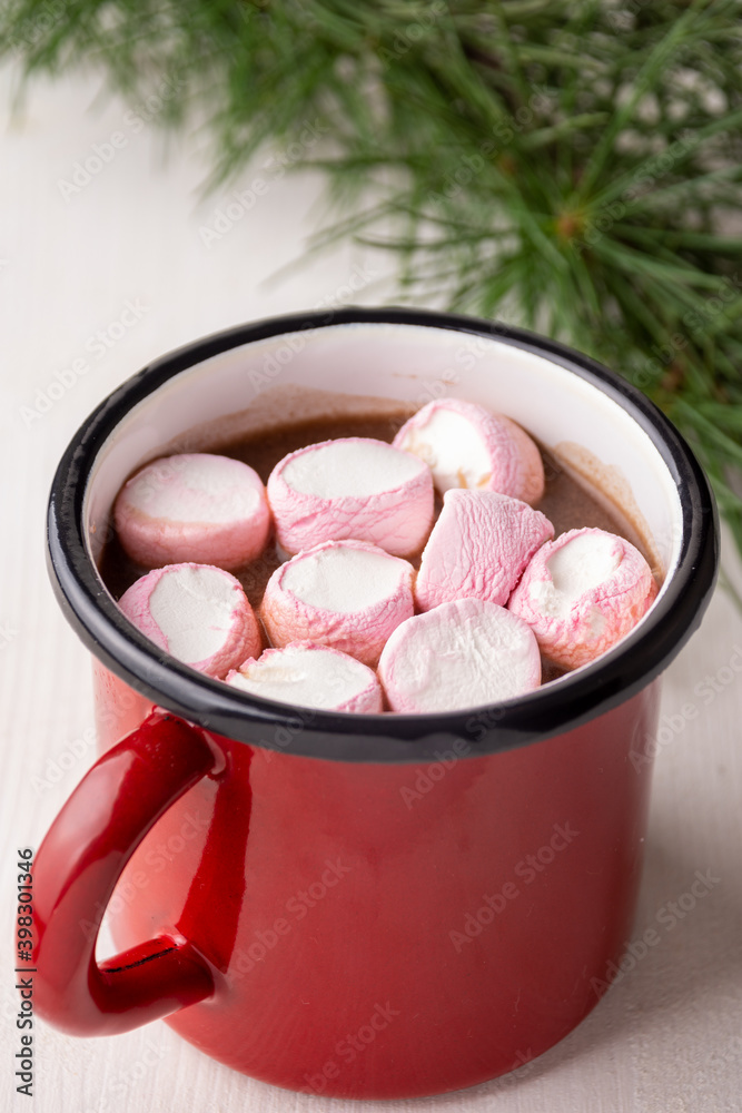 Aerial view of red mug with pink marshmallow and chocolate on white wooden table with green branch, vertical, with copy space