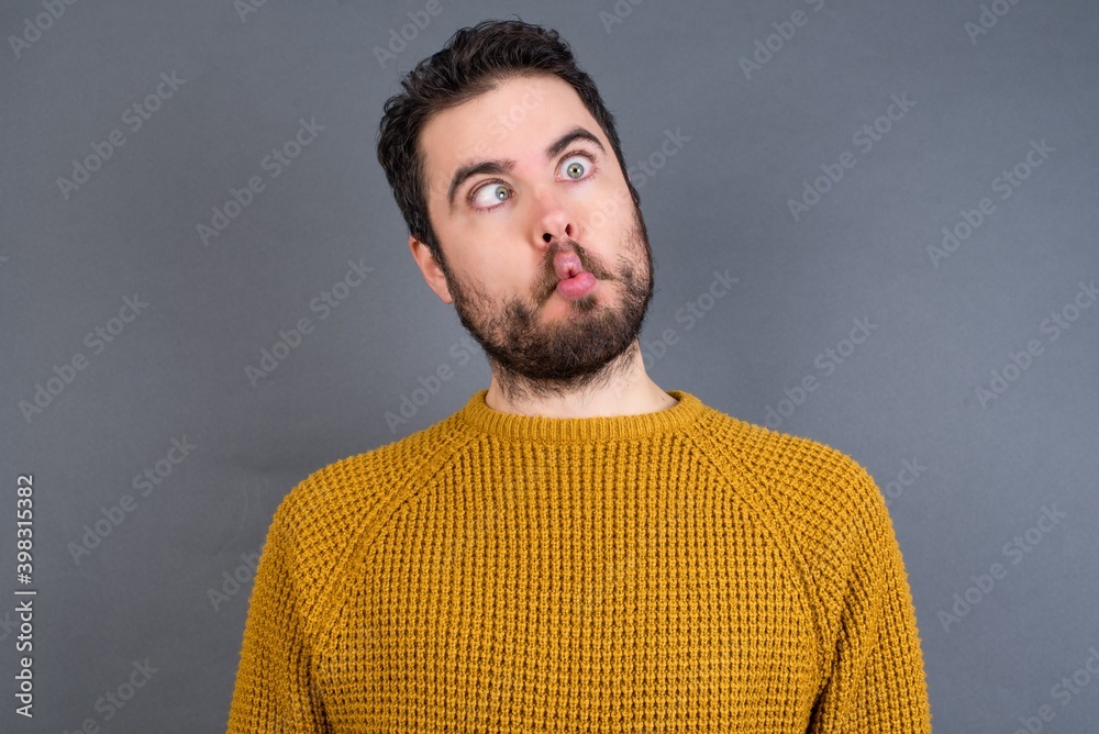 Obraz premium Young handsome Caucasian man wearing yellow sweater against gray wall making fish face with lips, crazy and comical gesture. Funny expression.