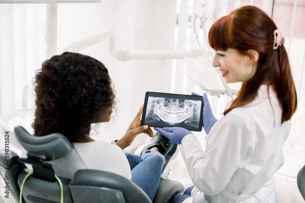 Young african woman patient sitting in dentistry chair and looking on the panoramic radiography. Pretty woman doctor explains the ways of treatment or shows good results of orthodontic treatment Young african woman patient sitting in dentistry chair and looking on the panoramic radiography. Pretty woman doctor explains the ways of treatment or shows good results of orthodontic treatment