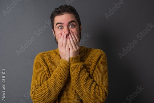 Vivacious Young handsome Caucasian man wearing yellow sweater against gray wall, giggles joyfully, covers mouth, has fun alone, has natural laughter, hears positive story or funny anecdote