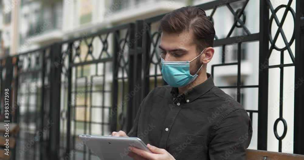 Young man in medical protective mask sits on the bench and uses tablet computer Young man in medical protective mask sits on the bench and uses tablet computer