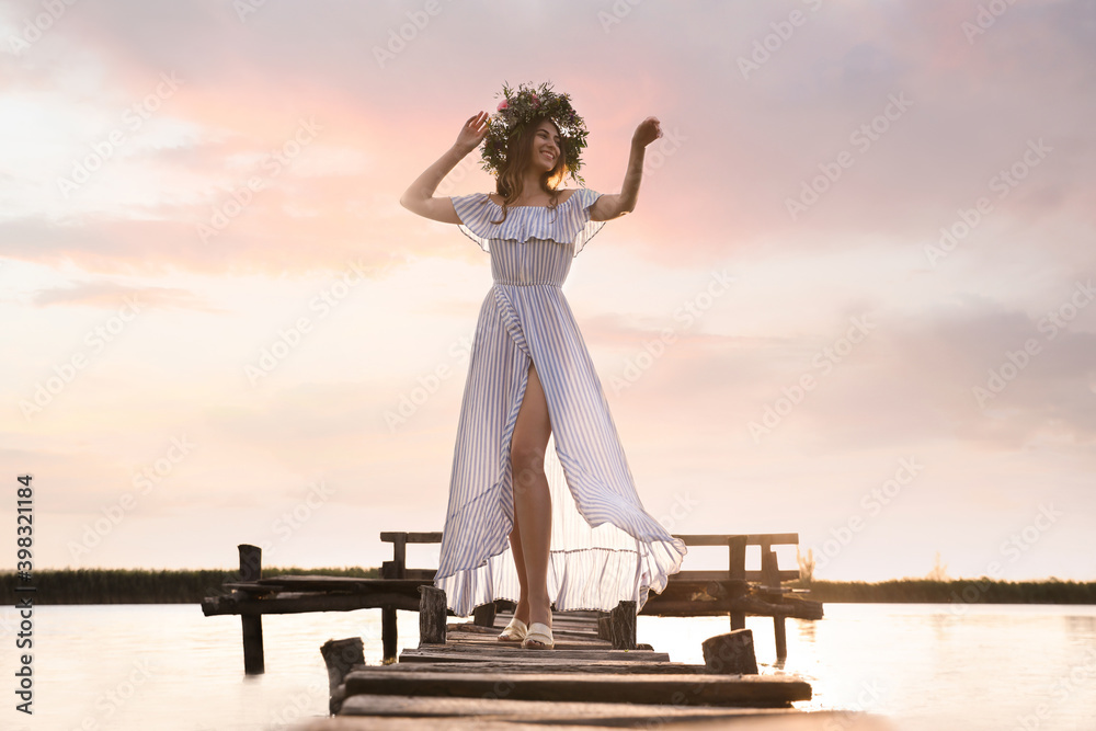 Fototapeta premium Young woman wearing wreath made of beautiful flowers on pier near river