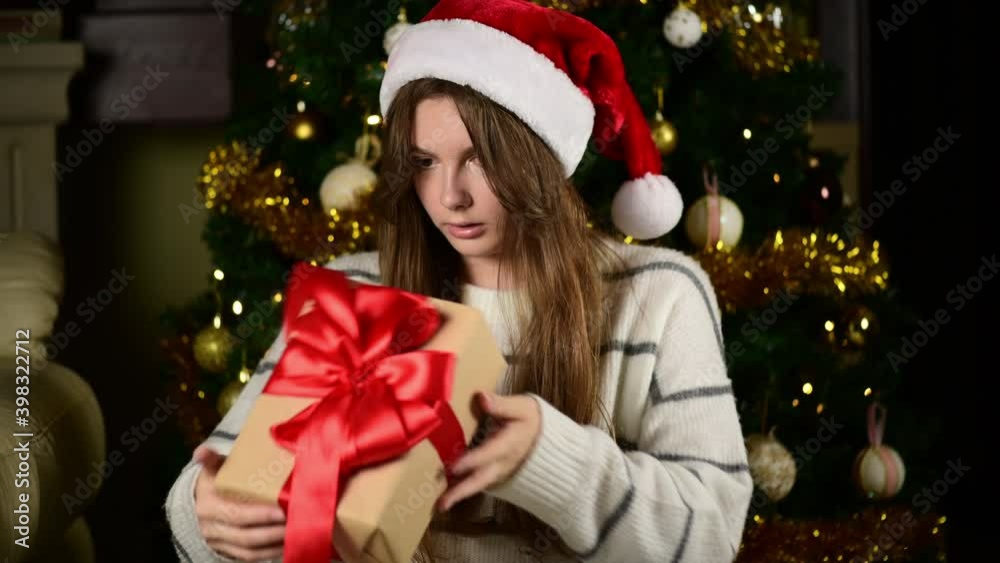 A teen girl sitting on the floor near the Christmas tree open gift. Happy young woman unwrapping Christmas present looking at box. Christmas spirit. She is surprised, rejoices at the surprise received
