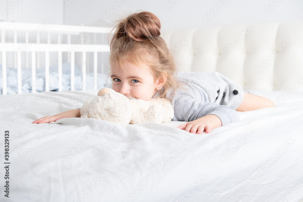 little girl in grey turtleneck with teddy bear laying on white bedding smiling.
