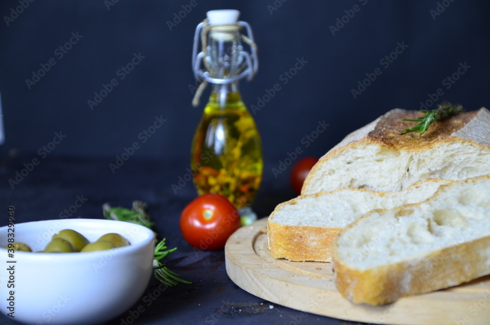 Italian ciabatta bread cut in slices on wooden chopping board with herbs, rosemary garlic and olives over dark grunge backdrop, top view