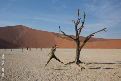 A girl tourist in a baseball cap and hiking clothes is jumping next to the dead trees in Dead Vlei against the background of a dune. Namibia