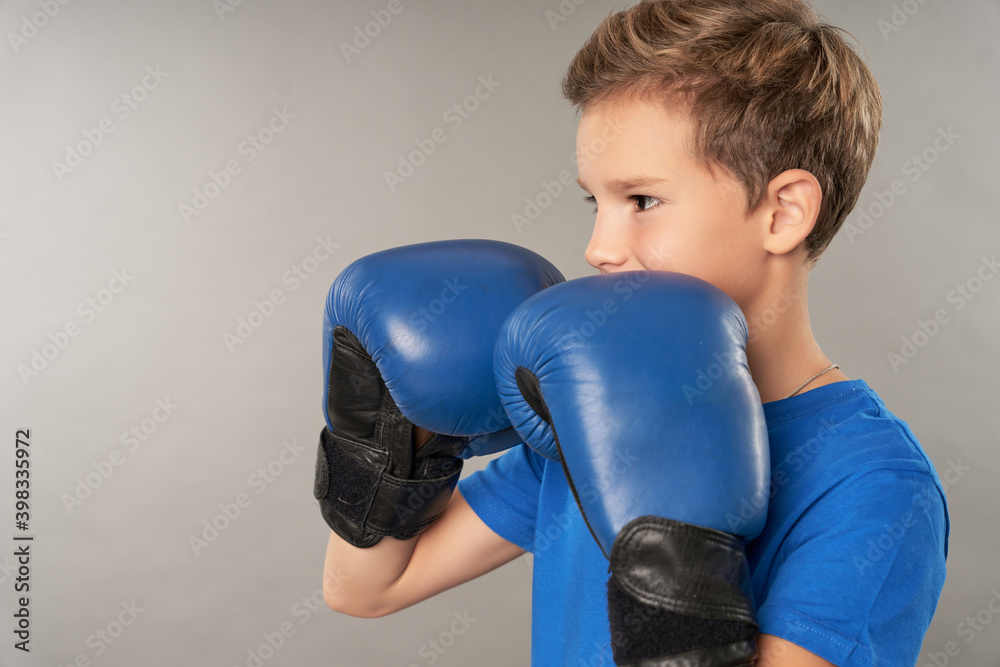 Cute male child in boxing gloves standing against gray background