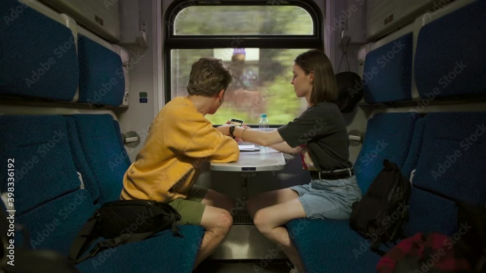 Young couple rides in a train compartment and a woman makes a photo of a young man on a smartphone. Guy and woman enjoy looking at smartphone in train compartment. Young couple rides in a train compartment and a woman makes a photo of a young man on a smartphone. Guy and woman enjoy looking at smartphone in train compartment.