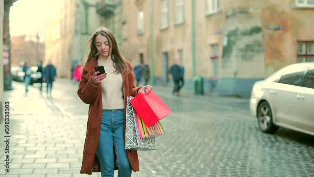 Young woman in a coat goes shopping after a sale on the street with a phone in her hands. Happy attractive girl with gift bags walks around the city on an urban outdoors background. Christmas presents Young woman in a coat goes shopping after a sale on the street with a phone in her hands. Happy attractive girl with gift bags walks around the city on an urban outdoors background. Christmas presents