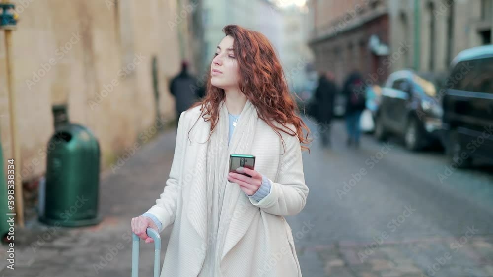 portrait young female tourist with a suitcase on wheels walking alone a street old town with a phone in her hands looking around. Woman traveling business in a coat visits city enjoys the application portrait young female tourist with a suitcase on wheels walking alone a street old town with a phone in her hands looking around. Woman traveling business in a coat visits city enjoys the application