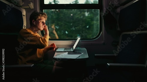 young man rides a train compartment in the evening and uses a laptop with wireless headphones. Freelancer working in traveling on laptop, riding train.