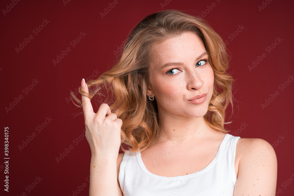 Blond woman in white tank top twists a curl on her finger on red background. Blond woman in white tank top twists a curl on her finger on red background.